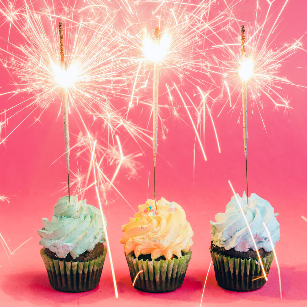 Drei bunte Cupcakes mit Creme-Topping und brennenden Wunderkerzen vor pinkem Hintergrund